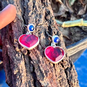 Peut inclure: Une paire de boucles d'oreilles en forme de cœur. Chaque boucle d'oreille présente une pierre en forme de cœur rouge profond facettée, sertie dans un cadre argenté, surmontée d'une petite pierre bleue. Fond texturé brun.