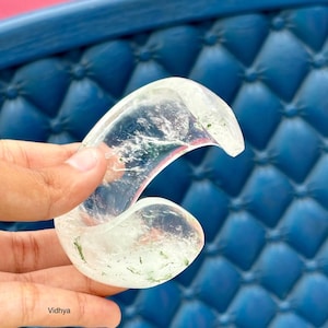 以下が含まれることがあります： 緑と白のインクルージョンが入った、波の形をした透明な水晶。青い背景に手の中に置かれている。