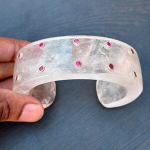 Könnte beinhalten: Transparente Quarz-Manschettenarmband mit eingebetteten roten Edelsteinen. Das Armband hat ein breites, gebogenes Design mit einer Reihe kleiner, runder roter Steine, die in den klaren Quarz eingesetzt sind. Das Armband wird von einer Hand vor blauem Hintergrund gehalten.