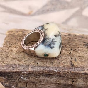 Puede incluir: Un anillo de plata con una banda decorativa con pequeños círculos. El anillo está engarzado en una piedra de color crema con patrones dendríticos negros y grises y una pequeña gema verde.