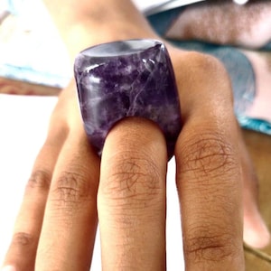 May include: A large, purple amethyst ring with a smooth, polished surface. The ring is worn on a finger and is a statement piece of jewelry.