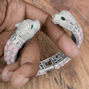 May include: A silver-toned bracelet featuring two panther heads encrusted with clear crystals and green eyes. The bracelet has a pink, oval-shaped stone design along the band. The bracelet is held in a person's hand.