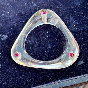 May include: A clear, triangular bangle with three red gemstones set into the metal.