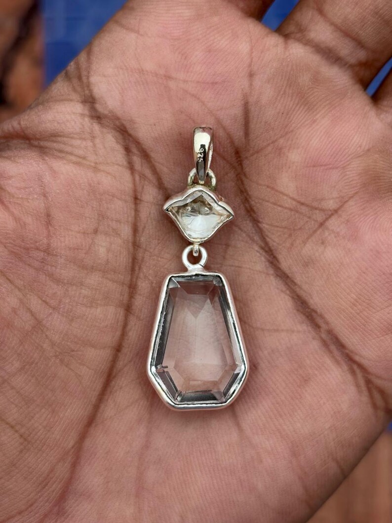 Pu&ograve; includere: Un pendente in argento con due pietre preziose chiare e sfaccettate. La pietra superiore &egrave; a forma di diamante, mentre quella inferiore &egrave; un esagono allungato pi&ugrave; grande. Il pendente &egrave; sospeso da un'ansa in argento.