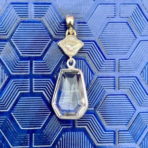 Pu&ograve; includere: Un pendente in argento con una gemma trasparente sfaccettata a forma di goccia e una gemma pi&ugrave; piccola a forma di diamante sopra. Il pendente &egrave; appeso a una cauzione color oro. Lo sfondo &egrave; una superficie blu strutturata.