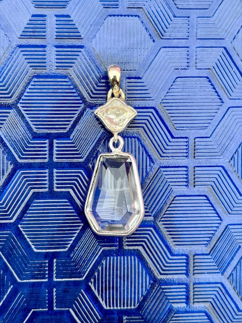 Pu&ograve; includere: Un pendente in argento con una gemma trasparente sfaccettata a forma di goccia e una gemma pi&ugrave; piccola a forma di diamante sopra. Il pendente &egrave; appeso a una cauzione color oro. Lo sfondo &egrave; una superficie blu strutturata.
