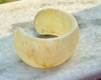 May include: A wide, curved cuff bracelet in a pale yellow hue. The bracelet has a smooth, polished surface with subtle variations in color, and a slight translucence. The word "Vidhya" is visible on the surface.