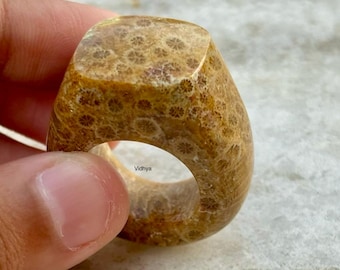 Puede incluir: Un anillo hecho de coral fosilizado. El anillo es de color marrón claro con un patrón de pequeños círculos blancos. El anillo es grueso y tiene una parte superior cuadrada.