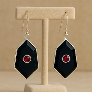 May include: A pair of geometric earrings with a black, diamond-shaped design and a small, red circular accent in the center. The earrings have a silver-colored border and are displayed on a beige jewelry stand.