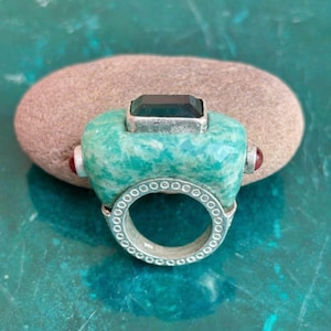 May include: A unique ring featuring a large, turquoise-colored stone with a dark rectangular gem at the top. The ring has a silver band with a circular design and small red stones on each side. The ring is set on a brown stone.