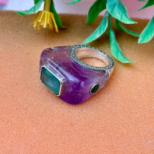 May include: A purple amethyst ring featuring a rectangular green stone and a small, round green stone. The silver band has a decorative pattern. The ring is set on a peach-coloured surface.