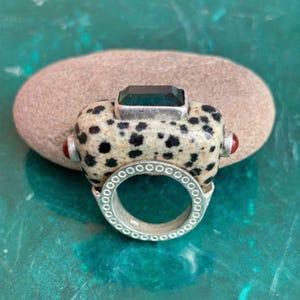Puede incluir: Un anillo único con una piedra moteada de color crema con manchas negras, una gema verde rectangular y dos pequeños detalles rojos. El anillo tiene una banda plateada con un patrón decorativo. Una pieza llamativa.