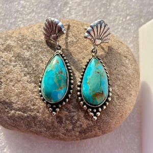May include: Silver-coloured earrings with turquoise teardrop-shaped stones. The earrings have a seashell design at the top and a beaded border around the turquoise stones. The earrings are displayed on a grey stone surface.