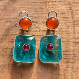 May include: A pair of silver earrings with a distinctive design. Each earring has a round orange stone at the top, a square teal stone in the middle, and a small red stone in the centre of the square. The earrings are on a wooden surface.