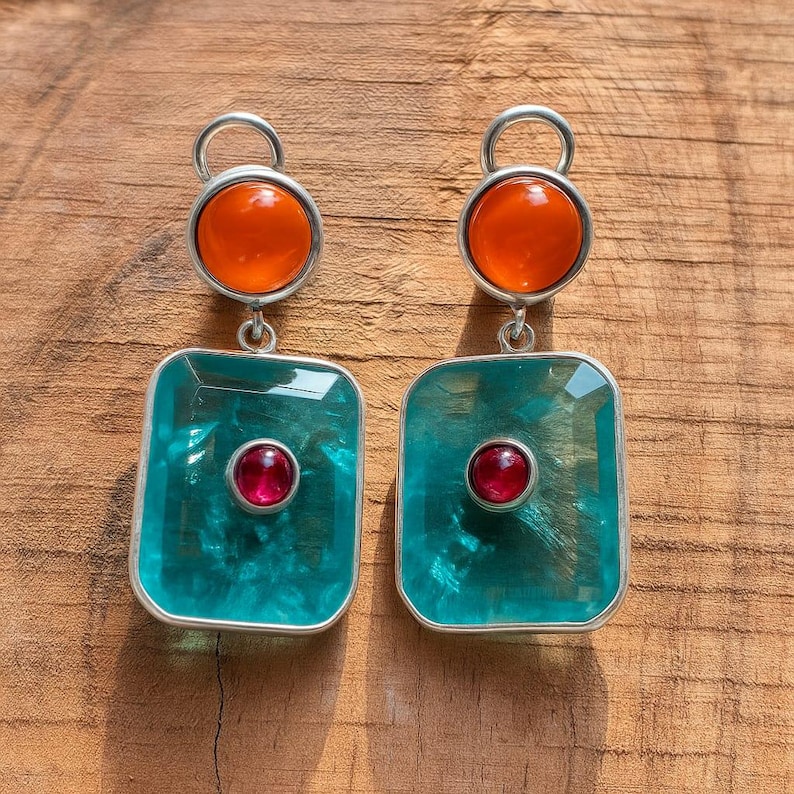 May include: A pair of silver earrings with a distinctive design. Each earring has a round orange stone at the top, a square teal stone in the middle, and a small red stone in the centre of the square. The earrings are on a wooden surface.