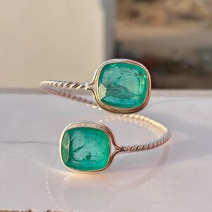 May include: A silver ring with two square, emerald-green gemstones. The ring's band has a twisted rope design. The gemstones are set in a square bezel setting, creating a modern look. The ring is displayed on a white surface.