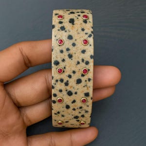 Könnte beinhalten: Ein breites, beigefarbenes Armband mit schwarzen Sprenkeln und eingelegten roten Edelsteinen. Die Edelsteine sind in goldfarbenen Fassungen gefasst, die gleichmäßig über die Oberfläche verteilt sind. Das Armband ist ein auffälliges Schmuckstück.