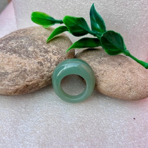 May include: A light green, translucent ring with a rounded top. The ring is made of a smooth, polished material. The ring is displayed on a white surface with two stones and a green plant.