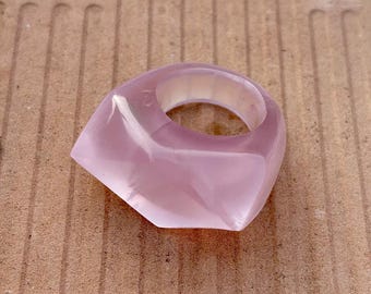 Può includere: Un anello rosa chiaro traslucido con un design geometrico. L'anello ha una forma quadrata con bordi arrotondati e una finitura liscia e lucida. È un accessorio di moda.