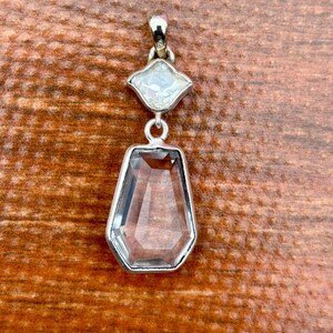 Pu&ograve; includere: Un pendente in argento con due pietre preziose trasparenti. La pietra superiore &egrave; una gemma bianca a forma di diamante, e quella inferiore &egrave; una gemma rettangolare, trasparente e sfaccettata. Il pendente &egrave; appeso a una cauzione d'argento.