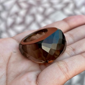 May include: A faceted, brown gemstone ring with a geometric design. The ring has a wide band and a large, rounded top. The gemstone is a translucent, smoky brown color. The ring is shown on a person's hand.