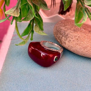 Puede incluir: Un anillo rojo oscuro en forma de corazón con una banda plateada con un diseño ornamentado. Una pequeña piedra preciosa verde está engastada en el lateral del anillo. El anillo se muestra sobre una superficie azul claro, con vegetación y una piedra en el fondo.