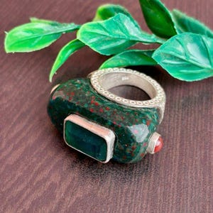 Op de afbeelding: Een unieke ring met een donkergroene steen met rode spikkels, een rechthoekige smaragdgeslepen groene steen en een kleine rode steen. De ring heeft een zilveren band met een decoratieve rand. Het is een sieraad.