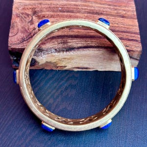 K&ouml;nnte beinhalten: Ein goldfarbenes Armreif-Armband mit sechs quadratischen, leuchtend blauen Edelsteinen. Das Armband hat ein filigranes Design mit kleinen, dekorativen Ausschnitten. Das Armband wird auf einer Holzoberfl&auml;che pr&auml;sentiert.