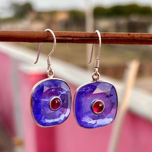 May include: A pair of silver earrings with a square, faceted, deep blue gemstone and a small, round, red gemstone in the center. The earrings hang from a silver hook.