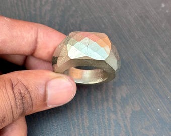 May include: A geometric ring with a faceted design, appearing metallic in a light colour. The ring's top surface is multi-faceted, creating a sparkling effect. The ring is held by a hand, showcasing its design and details.