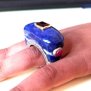 Könnte beinhalten: Ein Ring aus Lapislazuli mit einem Silberband und zwei roten Edelsteinen. Der Ring sitzt an einem Finger.