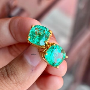 May include: A pair of gold earrings featuring large, square-shaped green gemstones. The gemstones have a sparkly, faceted finish.
