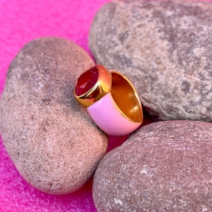 Puede incluir: Un anillo dorado con una gran gema roja facetada. El anillo tiene una banda de esmalte rosa y descansa entre varias piedras grises sobre un fondo rosa. El interior del anillo también es dorado.