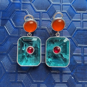 May include: A pair of silver earrings with a geometric design. Each earring has a round orange stone at the top, a large rectangular teal stone in the centre, and a small red stone in the middle of the teal stone.