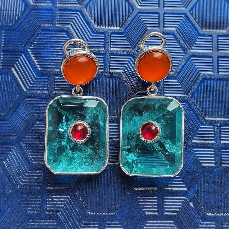 May include: A pair of silver earrings with a geometric design. Each earring has a round orange stone at the top, a large rectangular teal stone in the centre, and a small red stone in the middle of the teal stone.