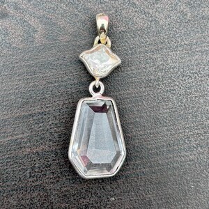 Pu&ograve; includere: Un pendente in argento con due pietre preziose chiare e sfaccettate. La pietra superiore ha una forma a diamante, e la pietra inferiore &egrave; un esagono allungato pi&ugrave; grande. Il pendente ha un design semplice ed elegante.