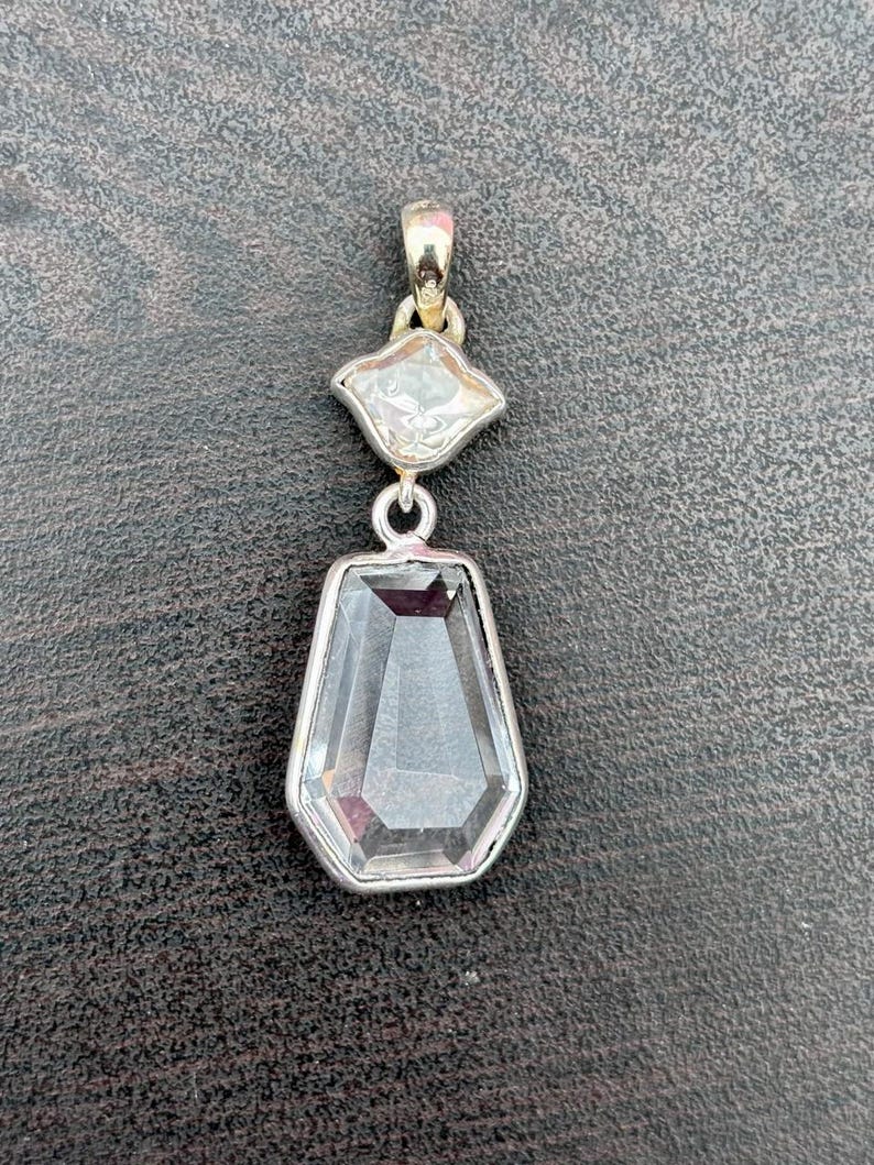 Pu&ograve; includere: Un pendente in argento con due pietre preziose chiare e sfaccettate. La pietra superiore ha una forma a diamante, e la pietra inferiore &egrave; un esagono allungato pi&ugrave; grande. Il pendente ha un design semplice ed elegante.