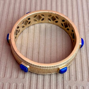 K&ouml;nnte beinhalten: Goldfarbenes Armreif-Armband mit geometrischen Aussparungen und eingelegten blauen Edelsteinen. Das Armband hat eine strukturierte Oberfl&auml;che und ein dekoratives Muster an den R&auml;ndern. Die Edelsteine sind rechteckig und gleichm&auml;&szlig;ig verteilt.