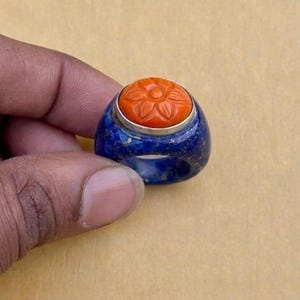 以下が含まれることがあります： 中央に彫刻されたオレンジ色の花が特徴の青いラピスラズリの指輪。指輪には花の周りに銀色のバンドがあり、手で持っています。ユニークなジュエリーです。