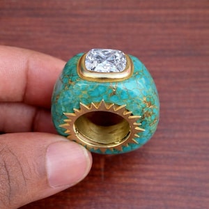 May include: A turquoise ring with a gold band and a large, square, clear gemstone. The ring has a unique design with a textured turquoise stone and a sunburst-like gold accent around the band.