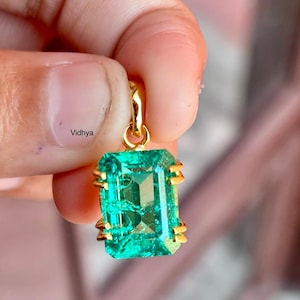 May include: A rectangular emerald-cut pendant featuring a bright green gemstone. The pendant is set in gold-coloured metal with decorative prongs. It hangs from a gold bail. The word "Vidhya" is printed on the image.