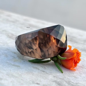May include: A geometric, faceted ring made of smoky brown gemstone. An orange flower with green stems and leaves sits beside the ring. The word "Vidhya" is visible in the background.