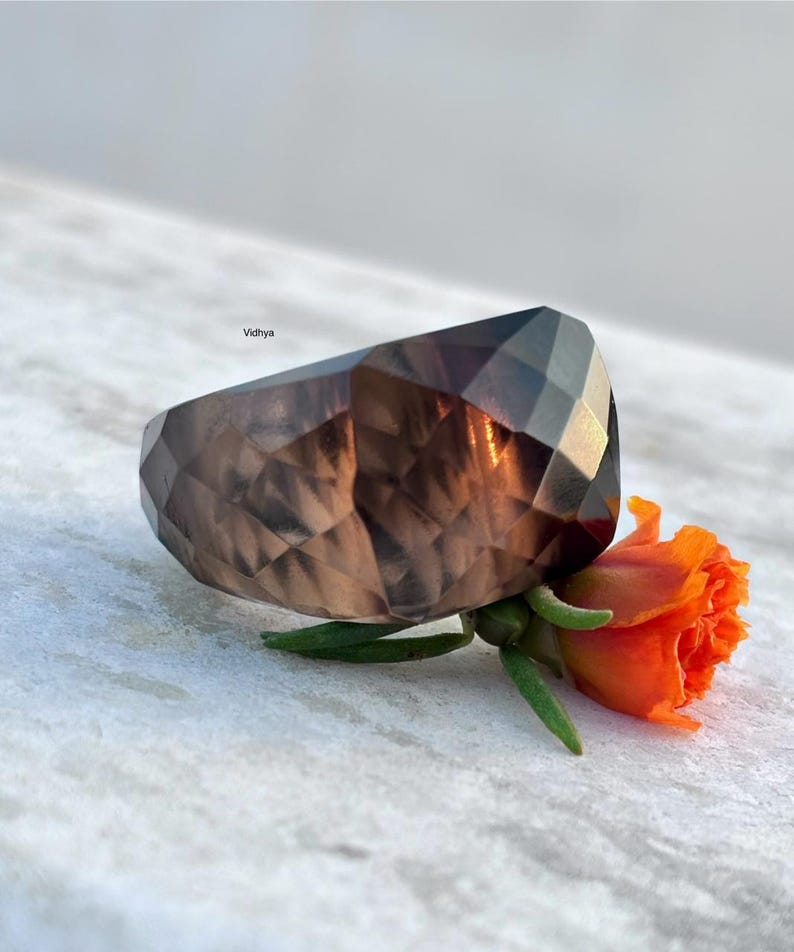 May include: A faceted, smoky brown gemstone ring with a geometric design. The ring is next to a small orange flower with green stems and leaves. The word "Vidhya" is in the background.
