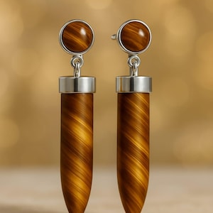 Op de afbeelding: Een paar oorhangers met een ronde, gepolijste, bruine steen aan de bovenkant, verbonden met een zilverkleurig metalen element. De oorbellen hebben een lange, cilindrische, bruine stenen hanger met een gestreept patroon. De achtergrond is wazig en neutraal.