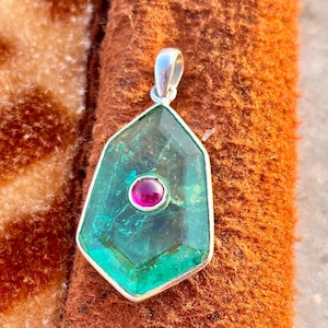 Puede incluir: Un colgante de piedra preciosa con una fianza plateada. El colgante presenta una piedra grande, facetada, de color verde azulado con una piedra más pequeña, redonda, de color magenta en el centro. El colgante se muestra sobre una superficie marrón texturizada.