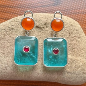 May include: Silver earrings with a striking design. Each earring has a round, orange gemstone at the top, linked to a rectangular, teal-coloured stone with a small, red gemstone in the centre. The earrings are presented on a textured, tan surface.