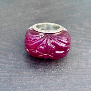 May include: A carved, deep magenta gemstone ring with a silver band. The gemstone has a floral design carved into its surface. The ring is set against a dark grey background.