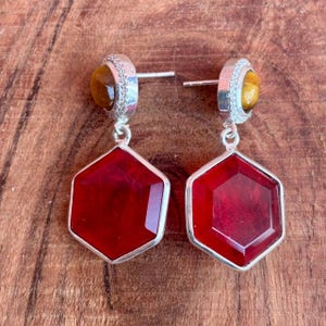 May include: A pair of silver earrings with a red, hexagonal gemstone pendant. The earrings have a small, round, amber-colored stone at the top, surrounded by small, clear stones. The earrings are displayed on a wooden surface.