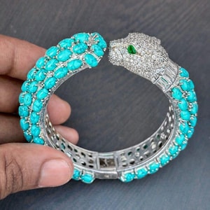 May include: A silver bracelet featuring a panther design. The bracelet is adorned with turquoise-colored stones and sparkling clear stones. The panther's eye is a green stone. The bracelet is a statement piece of jewelry.