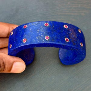 May include: A vibrant blue cuff bracelet with inlaid red gemstones. The bracelet has a curved design and is held by a hand. The gemstones are evenly spaced around the bracelet. The bracelet is made of a solid material.